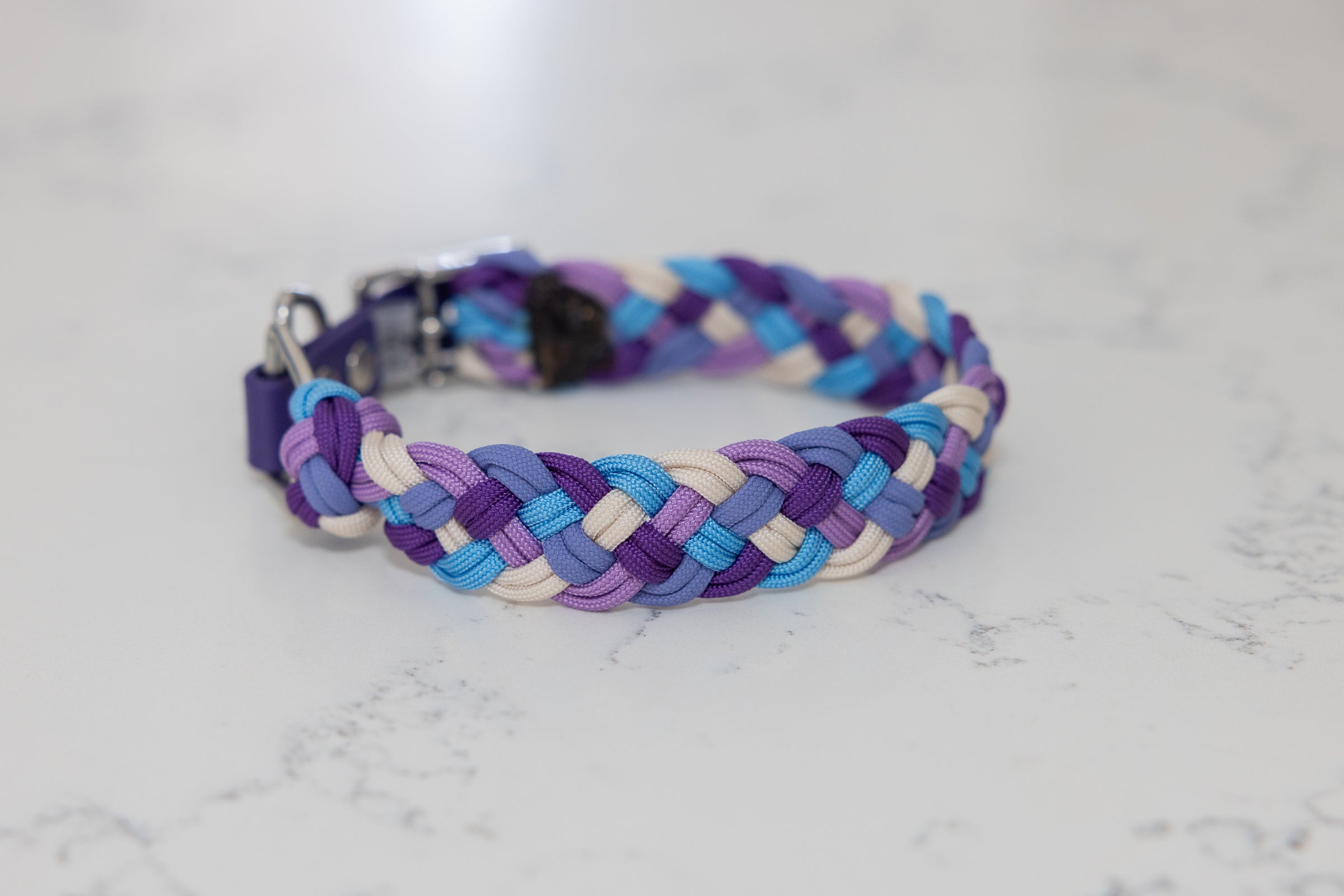 A multicolored paracord dog collar with purples, blue and cream strand displayed on a marble background.